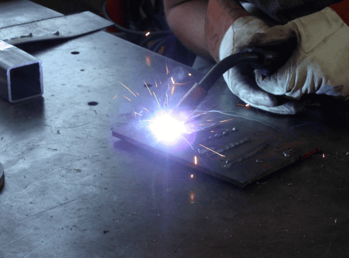 ชายใช้เครื่องเชื่อม MIG ในการทำงาน โดยมีประกายไฟและสวมอุปกรณ์ป้องกันความปลอดภัย