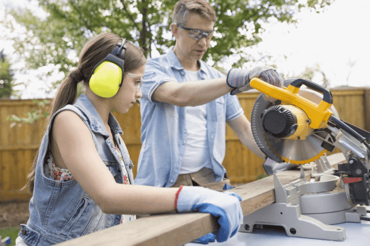 พ่อและลูกสาวกำลังทำงานร่วมกันในโรงงานไม้ โดยใช้เลื่อยมิตเตอร์ในการตัดไม้ ลูกสาวถือไม้แผ่นขณะที่พ่อกำลังใช้เลื่อย แสดงถึงการทำงานเป็นทีมและทักษะช่างไม้